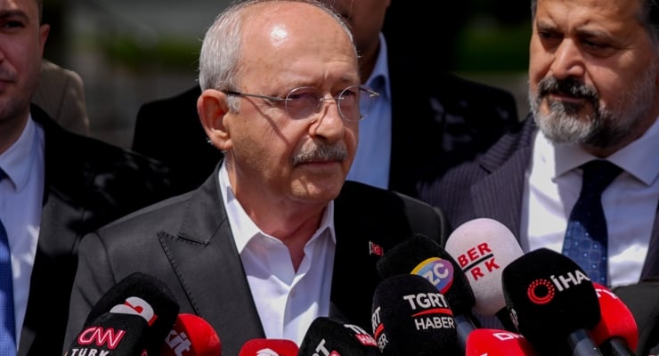 Older man with glasses in a dark suit and white shirt speaks at a press conference, surrounded by red microphones with Turkish media logos.