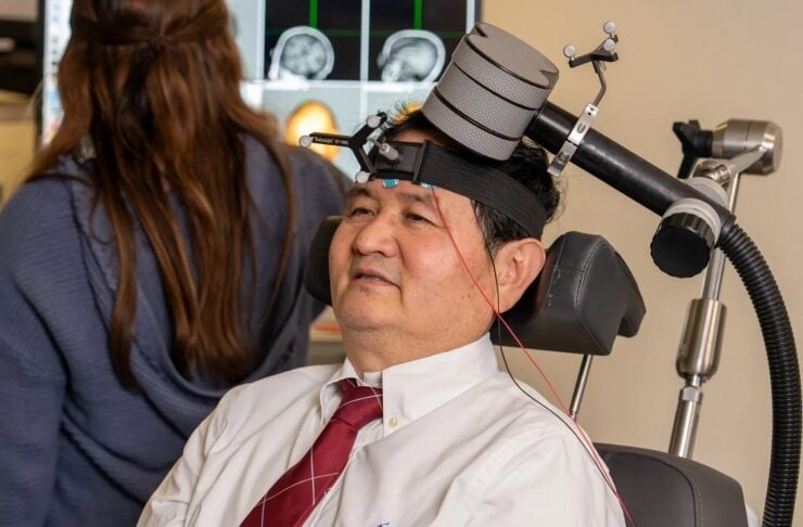 Man in a white shirt and red tie sits in a medical chair wearing a head-mounted device with straps and sensors during a neurological exam.