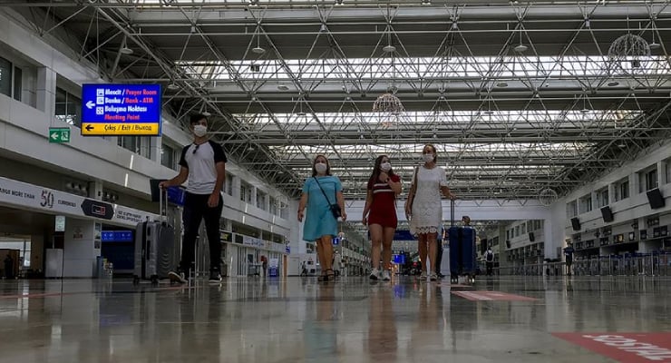 Four travelers wearing face masks walk through a spacious airport terminal with blue directional signs overhead.