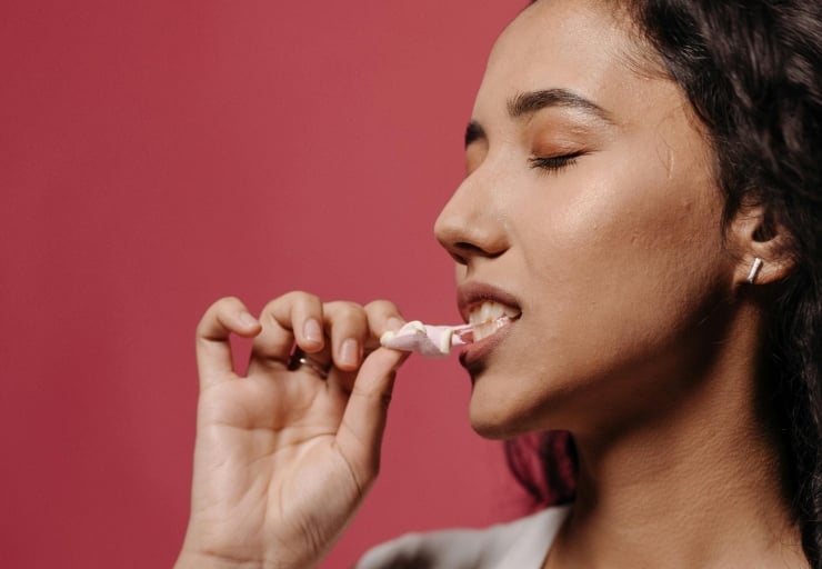 Close-up of a woman with eyes closed, biting a small pink-and-white candy held between her fingers against a pink background.