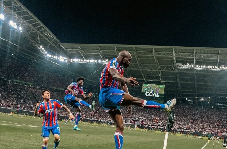 Soccer players in blue and red striped kits celebrate a goal near the corner flag at night, with stadium lights overhead.