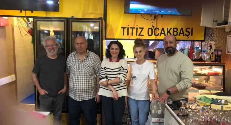Five adults stand side by side behind a counter in a kebab shop, smiling at the camera with a yellow sign overhead reading 'TITIZ OCAKBAŞI'.