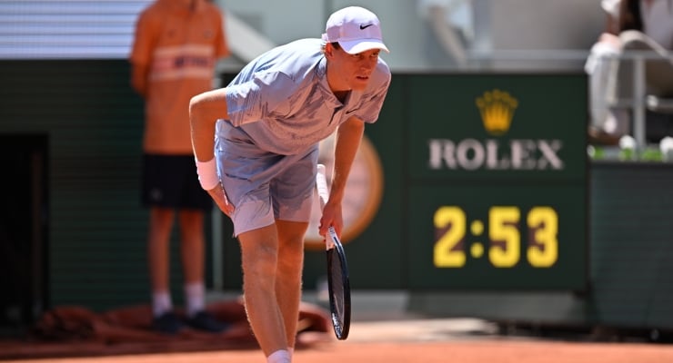 Tennis player bending down on a clay court, ready to hit with a racket, Rolex scoreboard showing 2:53 in the background.