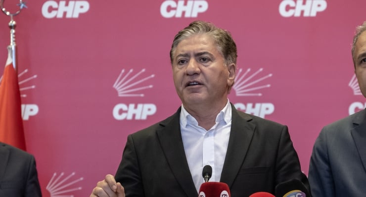 Man in a dark suit speaks at a press conference with CHP logos on a red backdrop behind him, microphones in front.