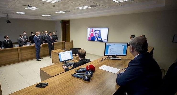 Officials seated at a long wooden table with microphones watching a video conference on a wall screen.