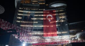Nighttime city scene showing a tall building draped with a large Turkish flag banner and portrait banners, with festive flags overhead.