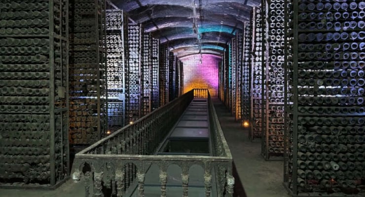 Underground wine cellar with tall racks of bottles lining a central corridor and colored lights at the far end.