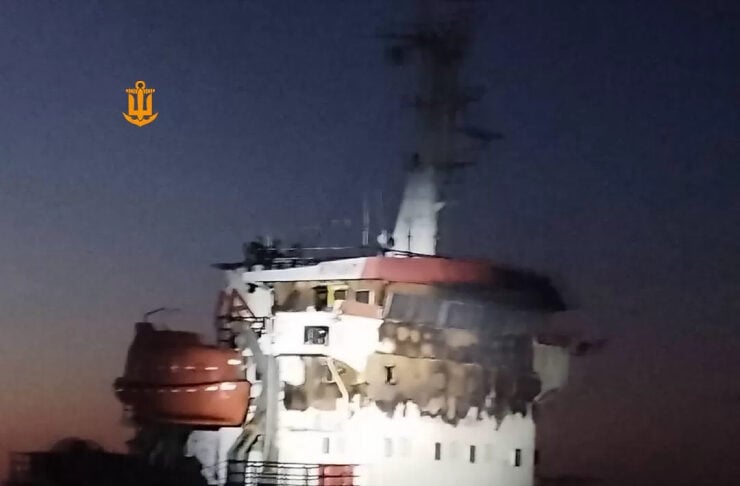 Nighttime view of a large tugboat with a white superstructure, blue hull, and an orange lifeboat on the side, illuminated against a darkening sky and choppy water.
