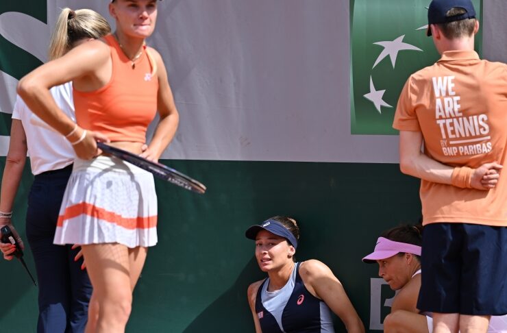 Tennis players on a clay court take a break; a female in navy suit sits with leg extended as others stand nearby, holding rackets.