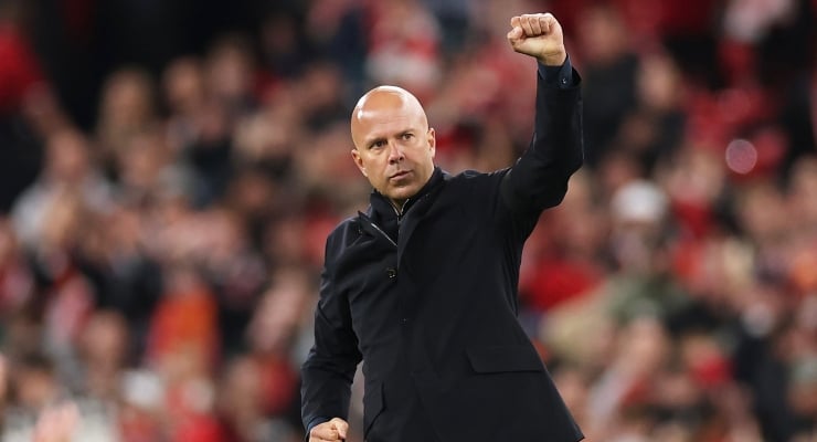 Bald man in a dark coat raises a clenched fist in celebration on the field, with a blurred stadium crowd behind him.