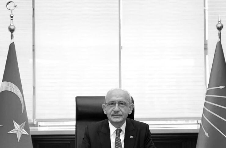 Government official seated at a large desk in a formal office, with flags on either side and documents on the desk.