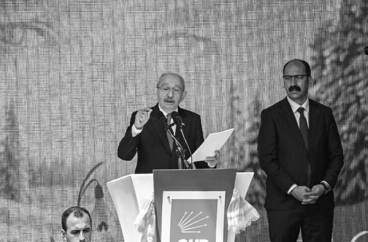 Older man in a suit speaks at a podium with CHP emblem, holding papers and gesturing to the crowd, flanked by another man in a suit on stage.