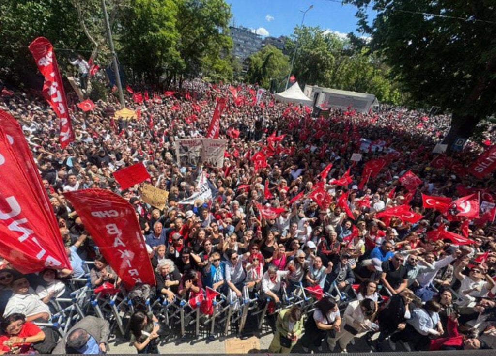 Large crowd gathers for a political protest, waving red flags and banners in a city square park area under sunny skies.