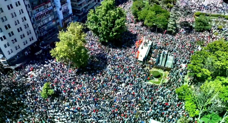 Aerial view of a dense crowd gathered in a city park surrounded by trees and tall buildings on the left side of the image, from above.