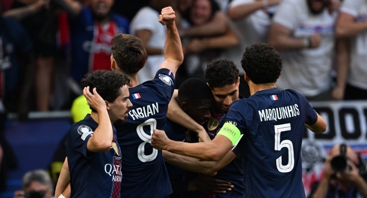 Group of Paris Saint-Germain players in navy jerseys celebrate together on the field, arms around each other after a goal, with a green captain's armband visible and cheering crowd in the background.