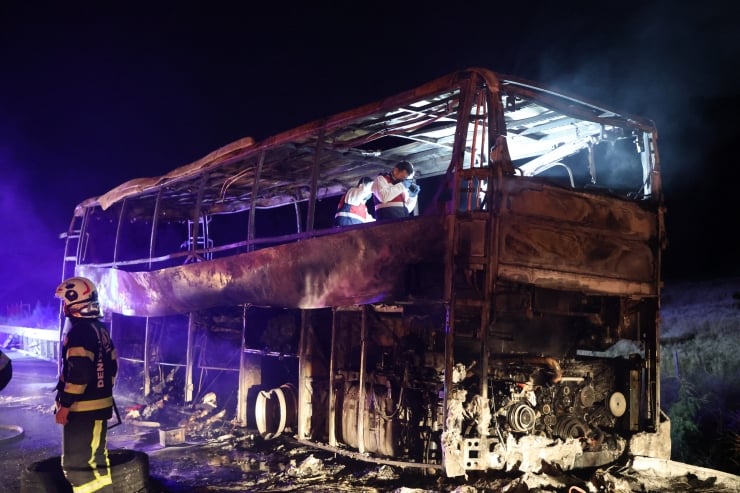 Charred, burned-out bus at night with firefighters inspecting the upper deck after a fire.