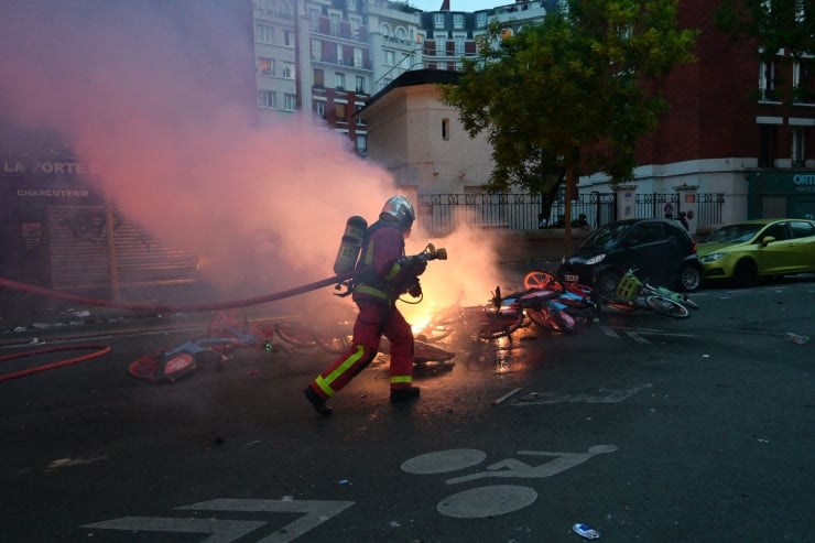 Firefighter in full gear approaches a street fire, with smoke and charred bicycles scattered on the road in an urban area.