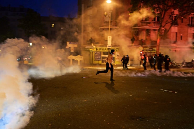 Shirtless man running through a smoke-filled street at night, with police visible in the distance near storefronts.