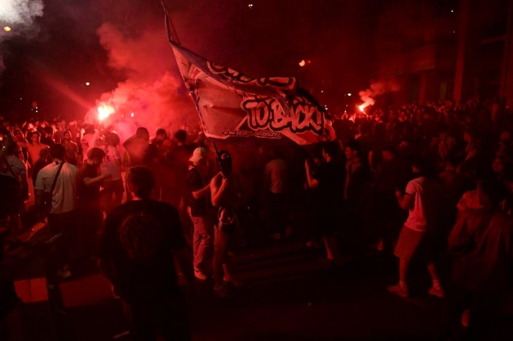 Crowd of protesters marching at night with red flares and a large banner raised above them.