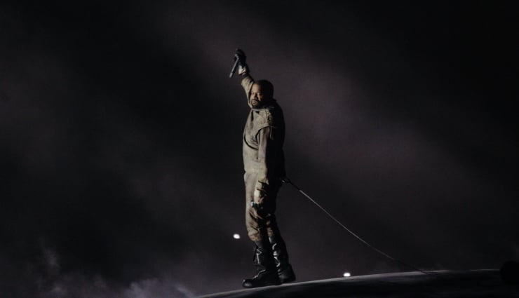 Man in protective gear stands on a vehicle at night, raising a knife above his head with a rope trailing behind him into the darkness.