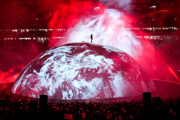 Person standing on top of a large globe-shaped stage prop amid red concert lights and smoke, surrounded by a cheering crowd at a stadium show.