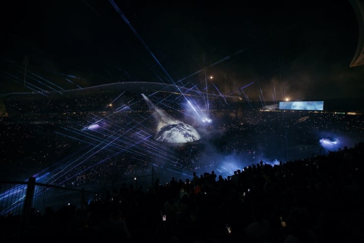 Nighttime stadium concert with a globe projection at center, surrounded by blue laser beams and fog, filled with cheering audience.