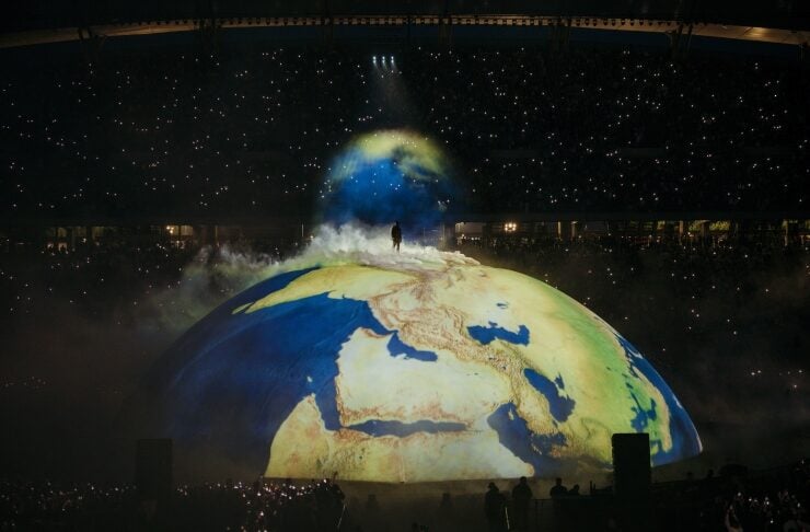 Person stands atop a large illuminated globe projection showing Africa and Europe, with fog, spotlights, and a cheering audience in a dark stadium behind.