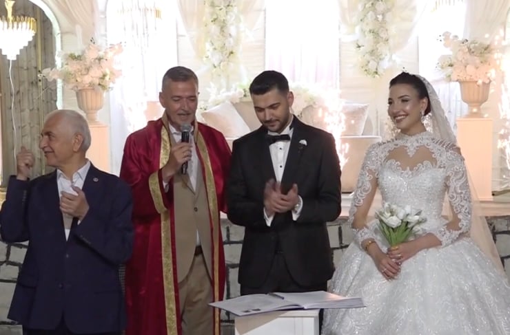 Bride in a lace wedding gown stands with the groom in a black suit as a officiant in red robes speaks into a microphone at the altar.