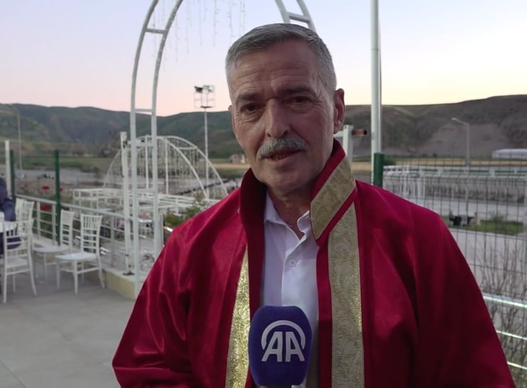 Man in a red traditional jacket being interviewed outdoors, holding a blue microphone with an AA logo, with hills and seating in the background.