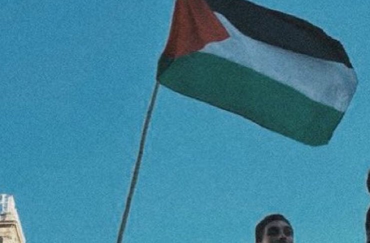 People on a balcony waving a Palestinian flag against a clear blue sky during a protest moment.