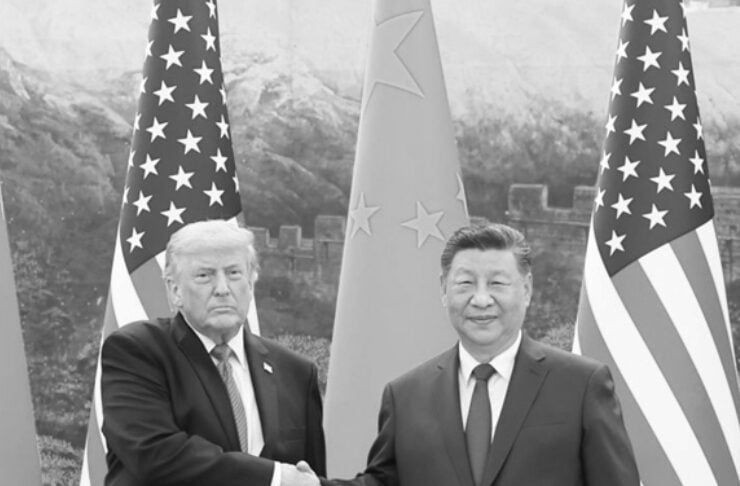 Black-and-white photo of two men in suits shaking hands, flanked by flags in a formal setting.