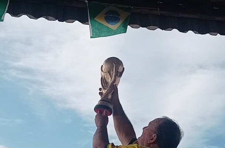 Man in a yellow shirt raises a trophy overhead in celebration, with Brazilian flag banners visible above.