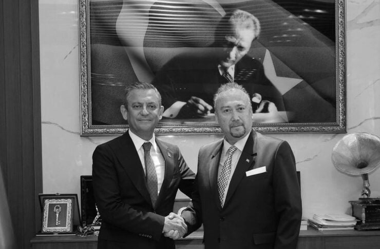 Two men in dark suits shake hands in a formal office, standing behind a desk with papers and a large framed portrait on the wall