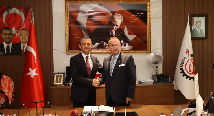 Two men in suits shake hands across a desk in a formal office, with Turkish flags and portraits in the background.