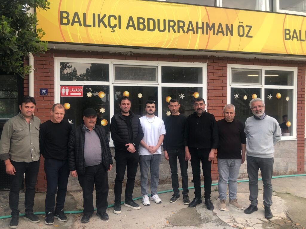 Nine men stand in a row outside a brick storefront with a yellow sign reading 'Balıkçı Abdurrahman Öz'.
