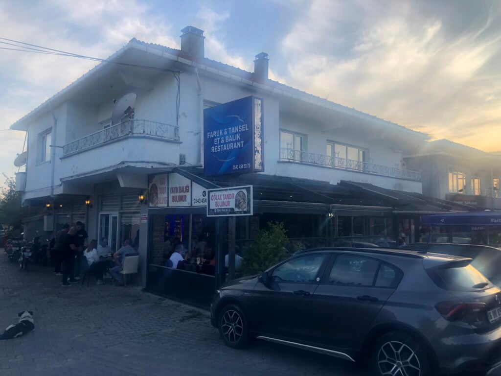 Two-story white building with a blue restaurant sign, outdoor seating along the street, diners at tables and a parked gray car nearby on a sunny evening.