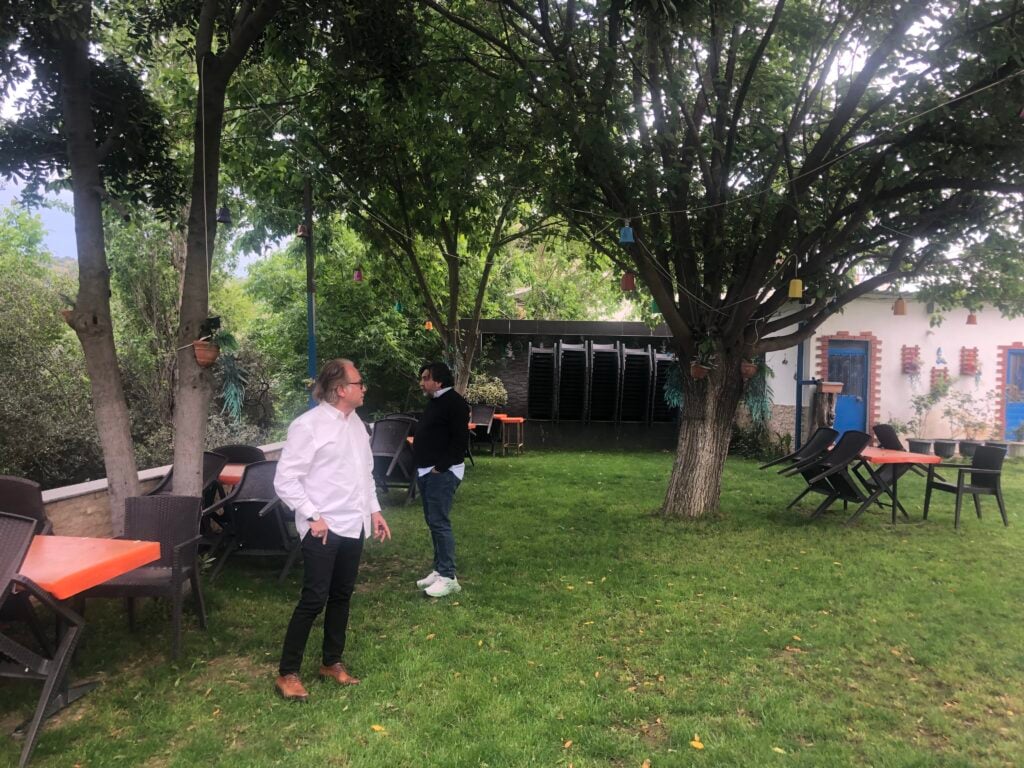 Two men in white and dark clothing stand and chat under a leafy tree in a garden with orange tables and stacked chairs nearby.