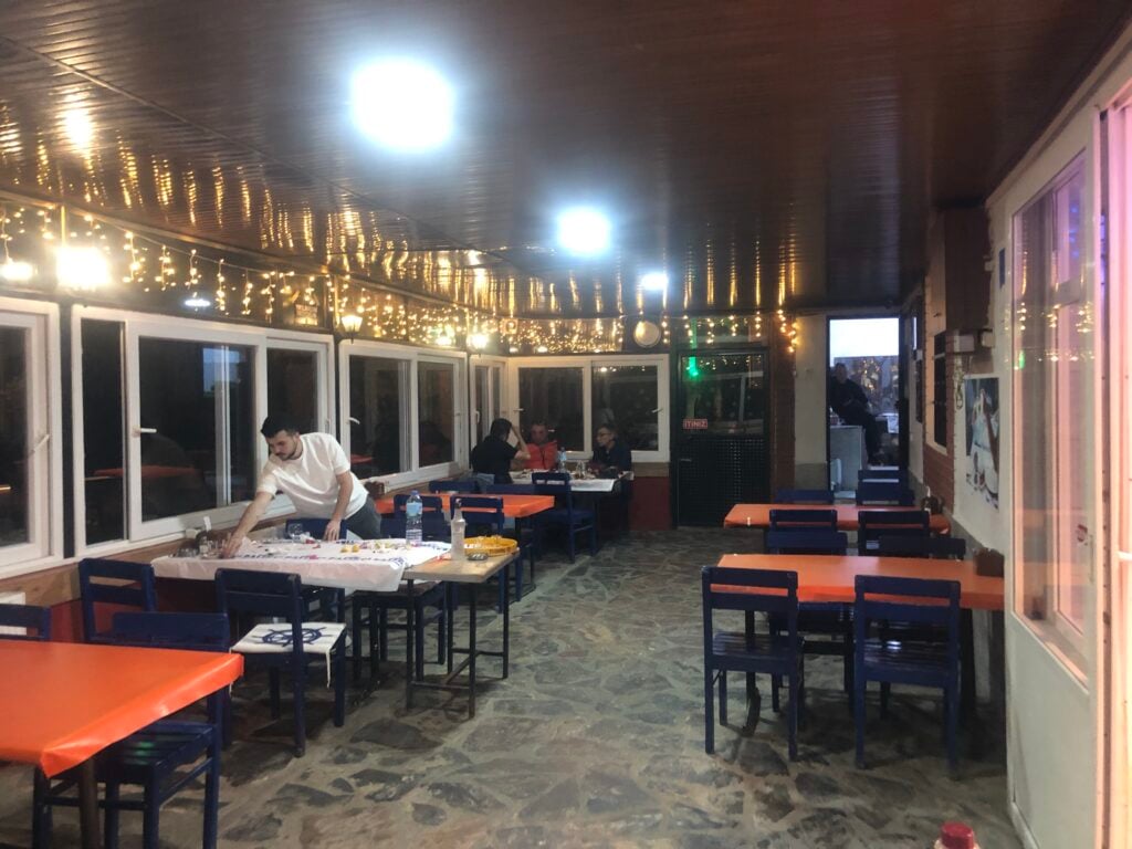 Indoor cafe scene with string fairy lights, orange tabletops and blue chairs, patrons dining by large windows.