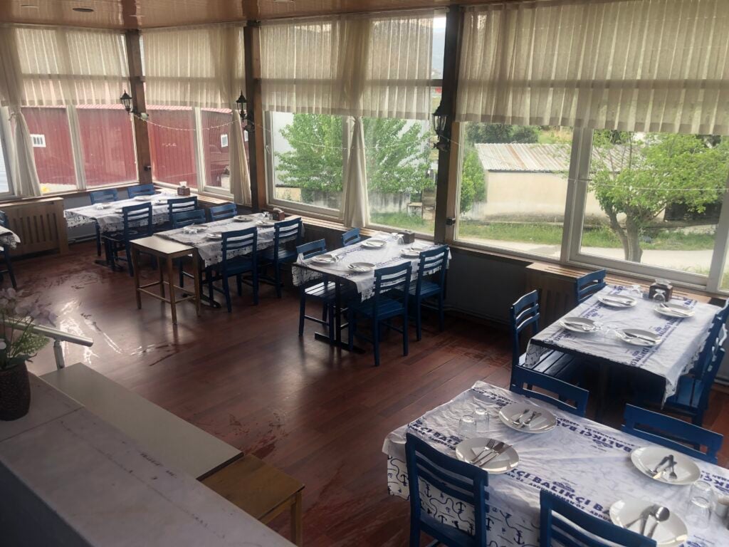 Sunlit dining room with long tables set for service, white patterned tablecloths, and blue chairs by large windows.