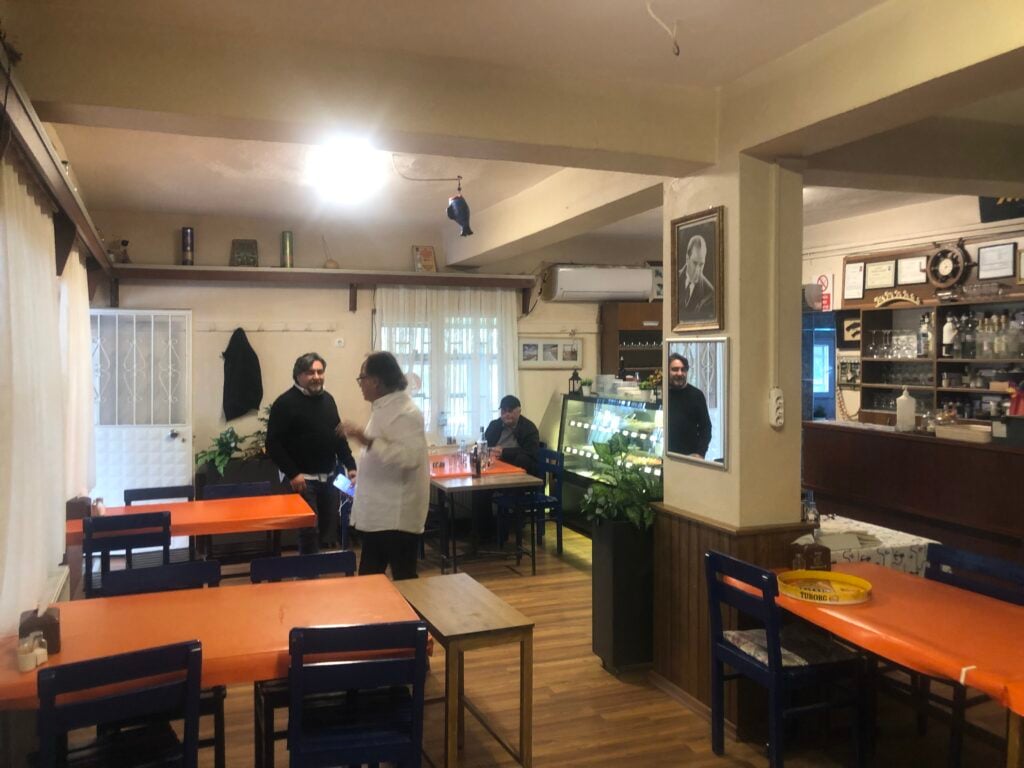 Cozy cafe interior with orange tables and blue chairs, several people standing and talking near a glass display counter. shelves and framed pictures line the walls behind them.