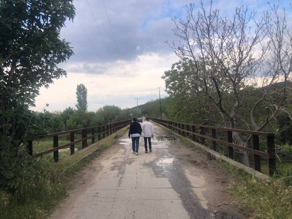 Two people walking away on a wet bridge path with railing, surrounded by trees under a cloudy sky.