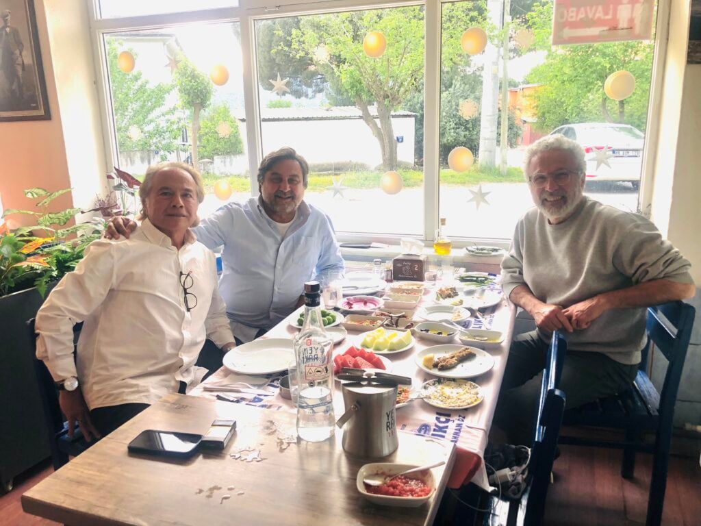 Three men sit at a sunny restaurant table with plates of food, drinks, and a watermelon slice visible, smiling at the camera.