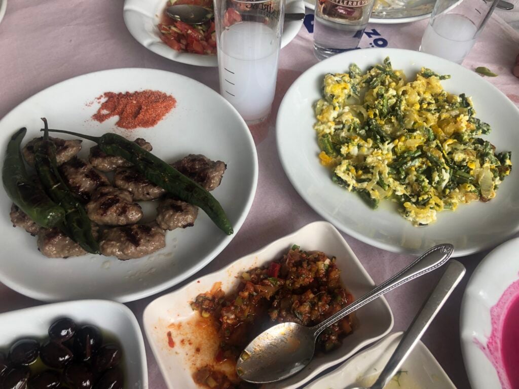 Dining table with several dishes: grilled meat and green peppers, scrambled eggs with greens, a tomato-based relish, and a bowl of black olives, with drinks in the background.