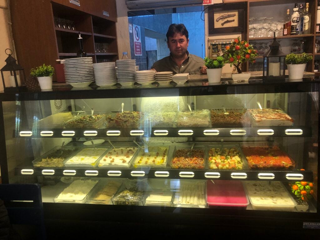 Man behind a glass deli case in a cafe, with stacked plates and small plants on top. Food trays line the shelves inside the case.