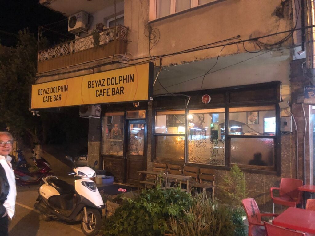 Night exterior of Beyaz Dolphin Cafe Bar with a yellow sign, warm-lit windows, outdoor seating, and a scooter parked nearby.