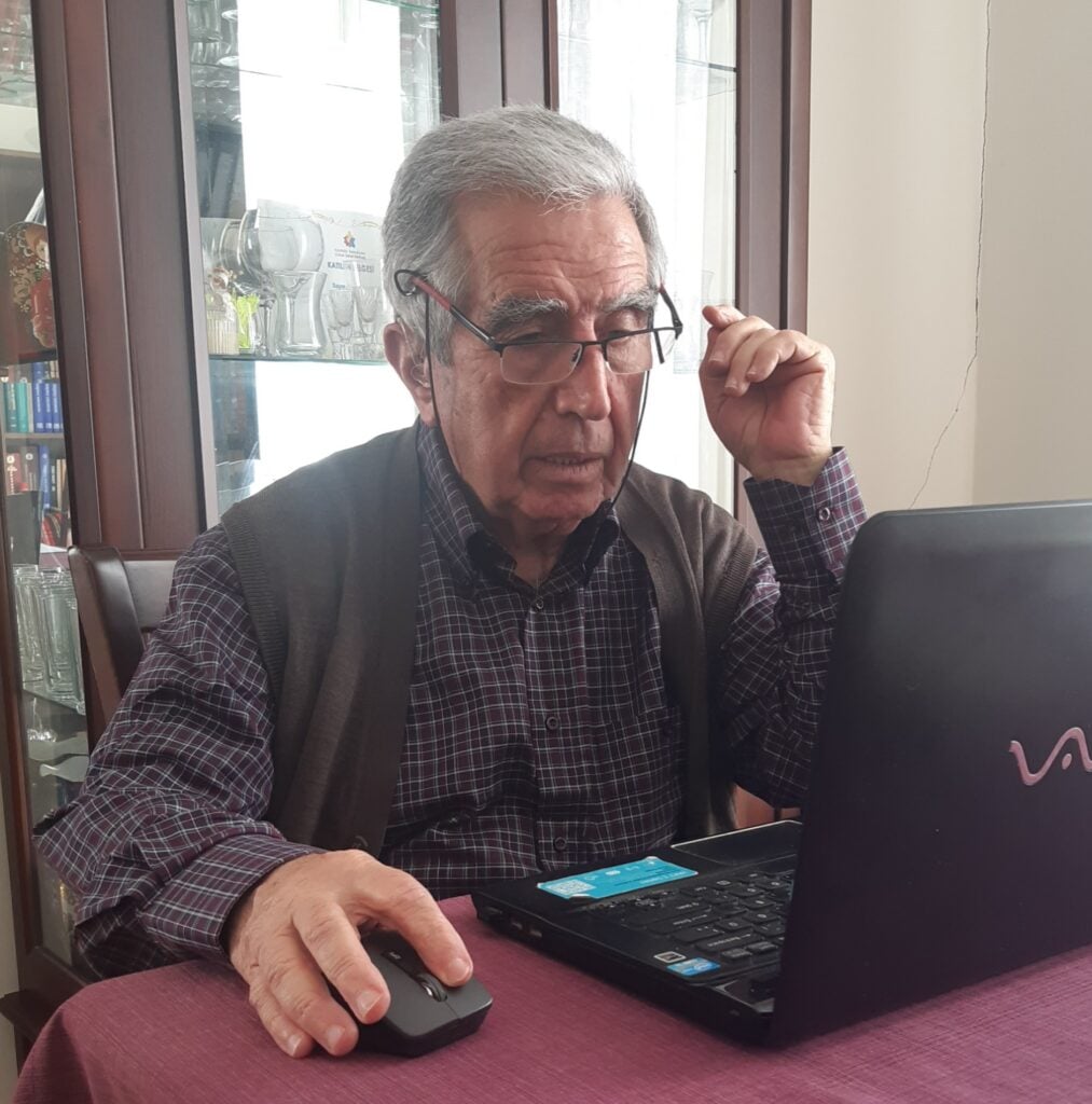 Older man with gray hair and glasses using a laptop, with a wireless mouse in his hand at a table indoors.