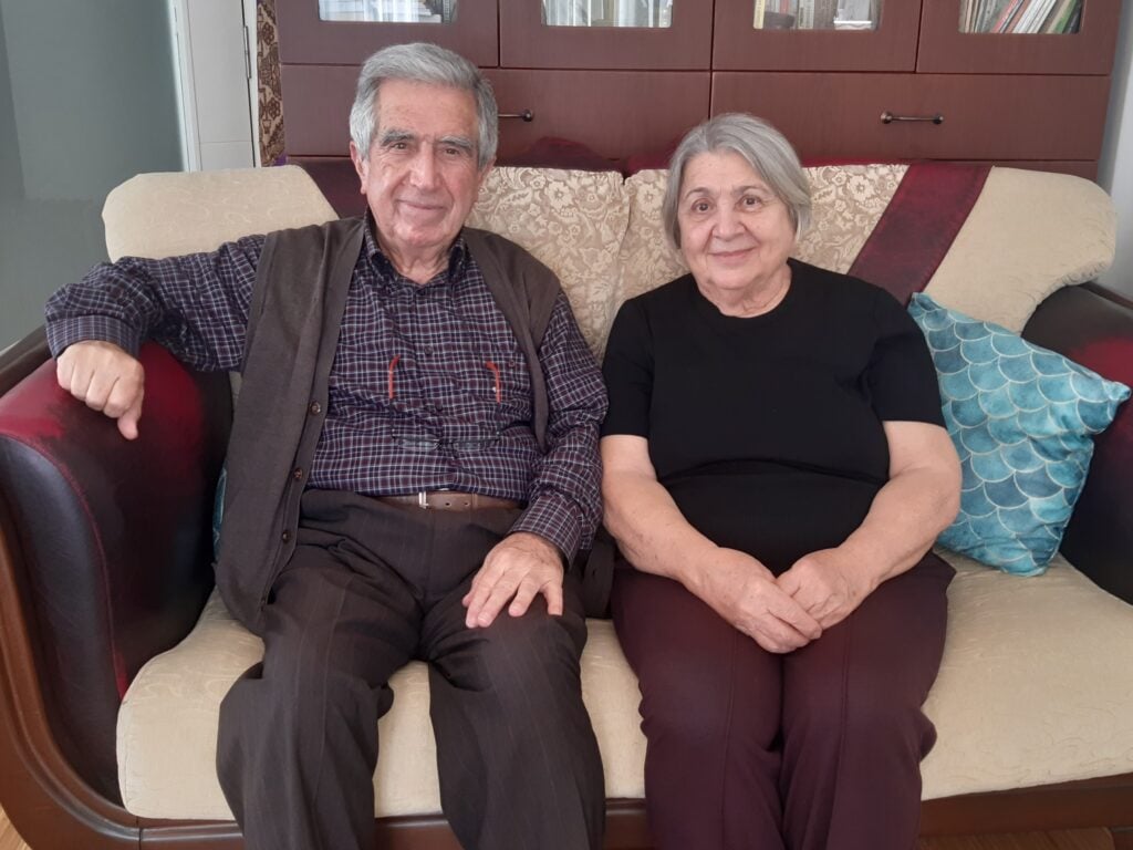 Elderly man and woman sit together on a beige sofa in a living room, smiling at the camera.
