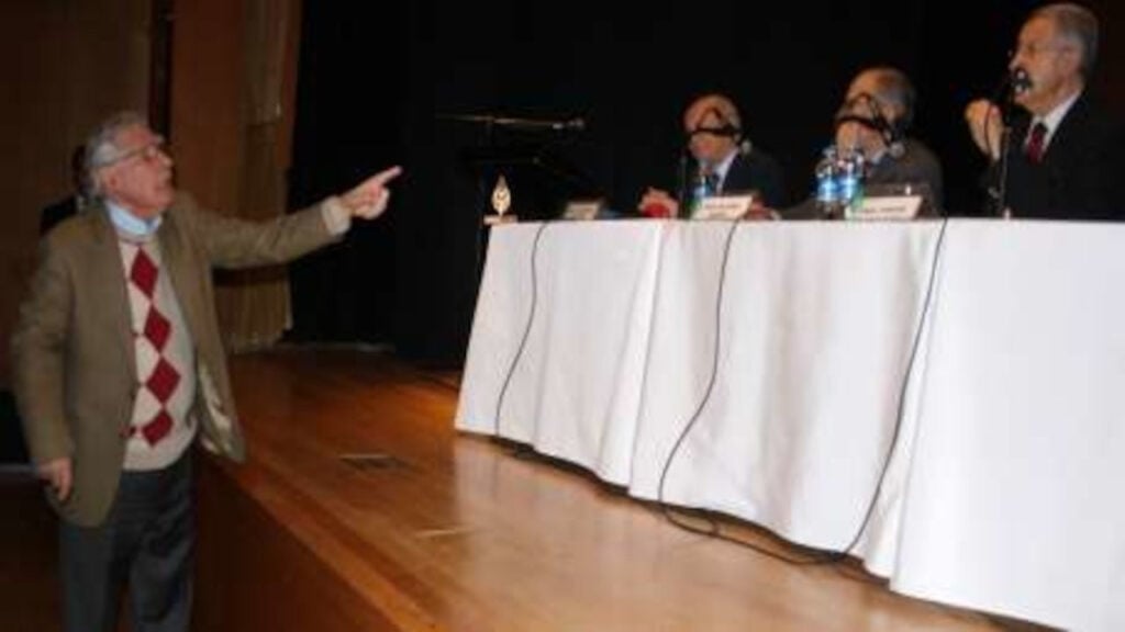 Older man in a blazer points toward a panel of speakers seated behind a long white table on a stage.