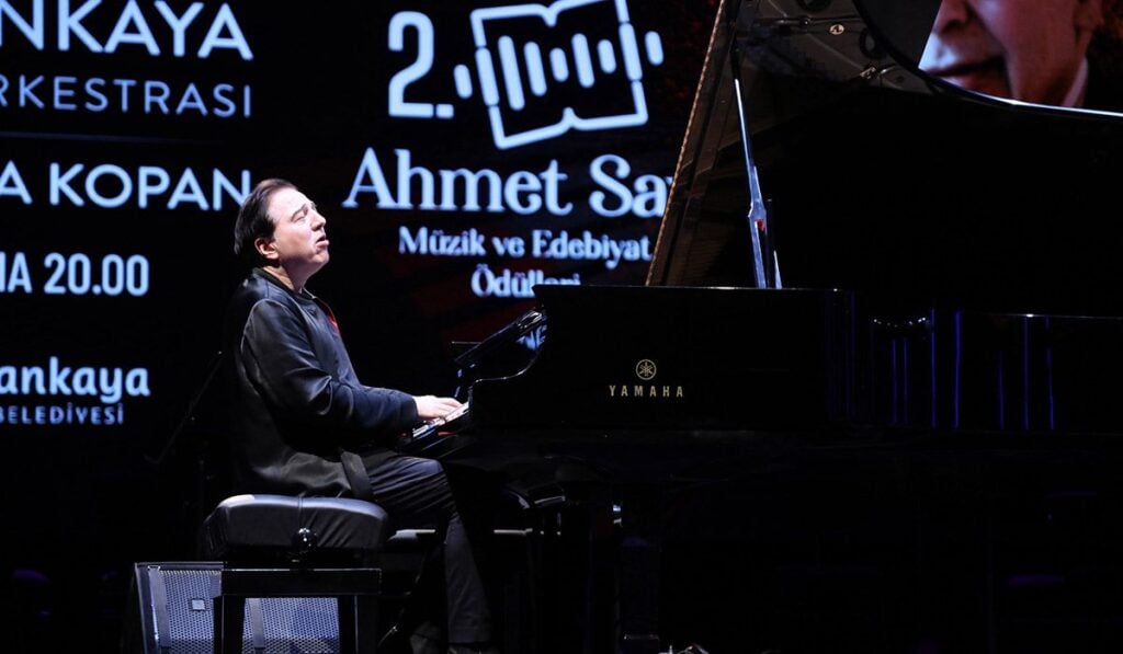 Male pianist seated at a Yamaha grand piano on a dark stage, illuminated backdrop with text and logos behind him, mid-performance.