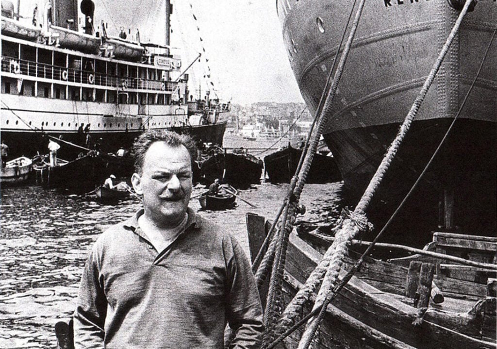 Man in a polo shirt stands on a dock with large ships and thick ropes behind him in a busy harbor (black-and-white photo).